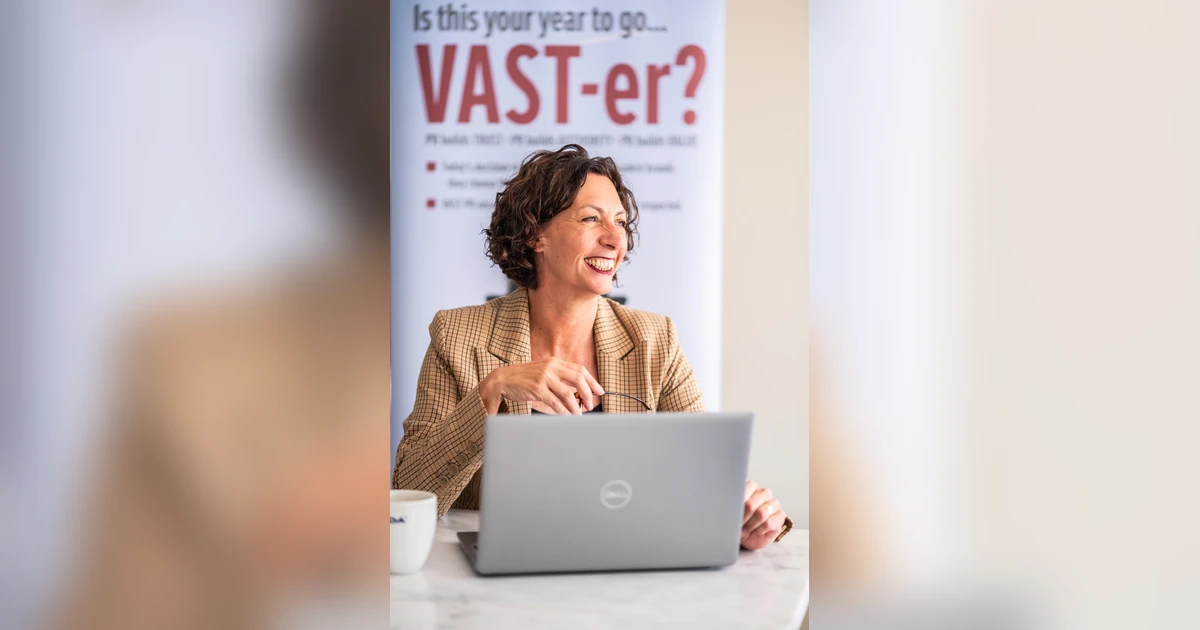 Woman in checked blazer smiling while using a laptop at a desk with a coffee cup and a business banner in the background