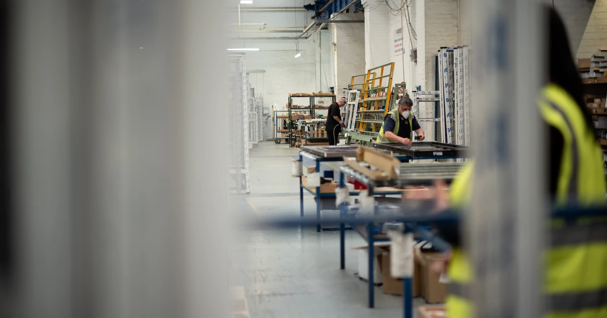 Three manufacturing workers wearing high-visibility clothing and masks working on assembly tables inside a factory production area.
