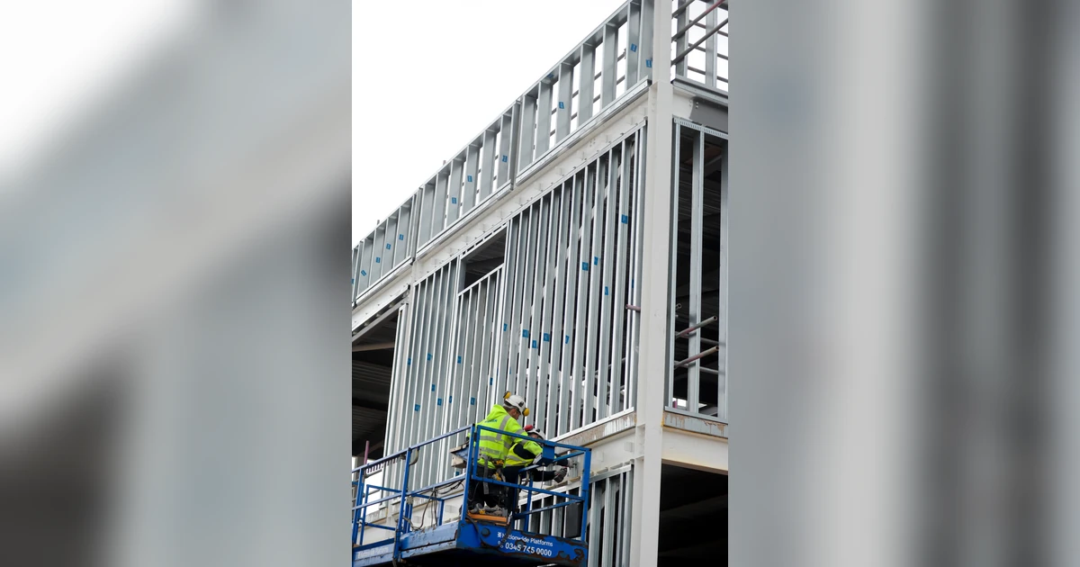 Two workers in high-visibility jackets on a lift working on the steel frame of a school building under construction