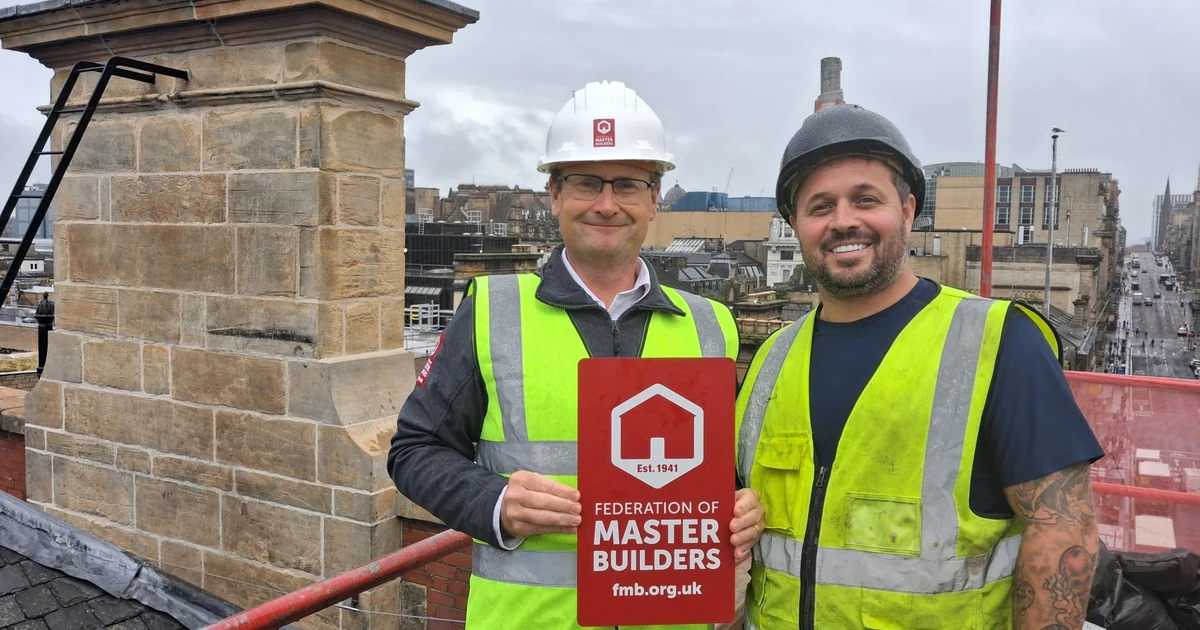 Two construction workers in safety vests and helmets on a rooftop with cityscape background, one holding a Federation of Master Builders sign.