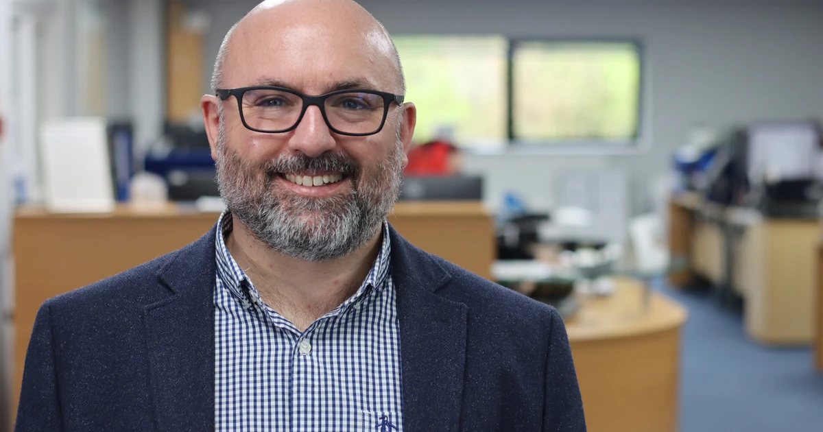Richard Hammond with glasses and beard standing in an office environment, smiling