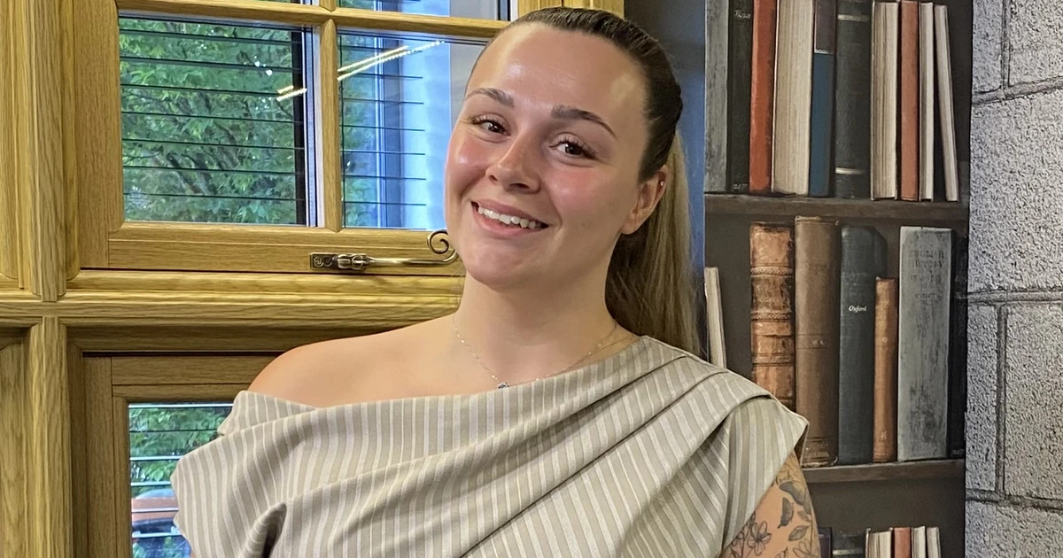 Alyssa Jones smiling, standing indoors next to a window with bookshelf wallpaper behind her