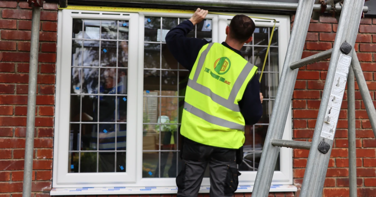 Installer wearing a high-visibility vest fitting a window into a brick house using a ladder