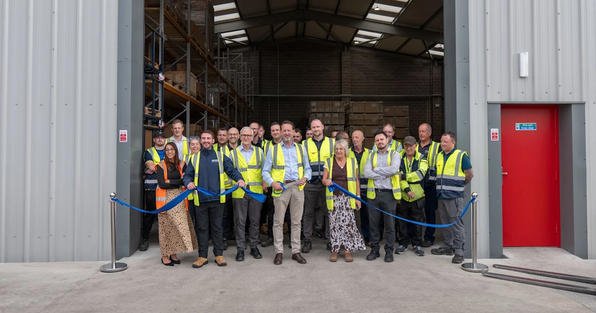 Group of Endurance employees in high-visibility vests standing at the entrance of the new Nexus warehouse building, holding a blue ribbon for opening event.