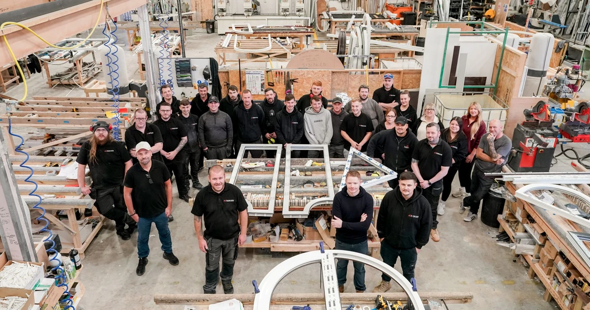 Twenty-six people from Premier Arches team standing in a large production facility with bespoke-shaped PVCu frames and manufacturing equipment.