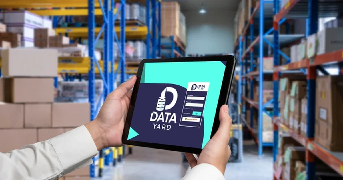 Hands holding a tablet displaying the Data Yard login screen in a warehouse with stacked boxes on shelves