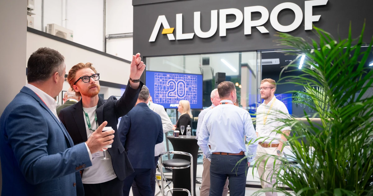 Group of business attendees mingling and conversing in front of an Aluprof UK showroom sign and a digital 20th anniversary display