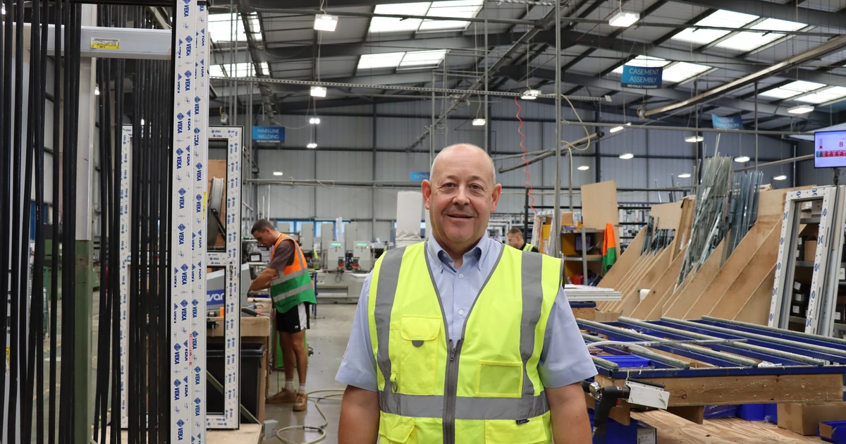 Man in a high-visibility vest smiling inside a busy window frame manufacturing factory with another worker in the background