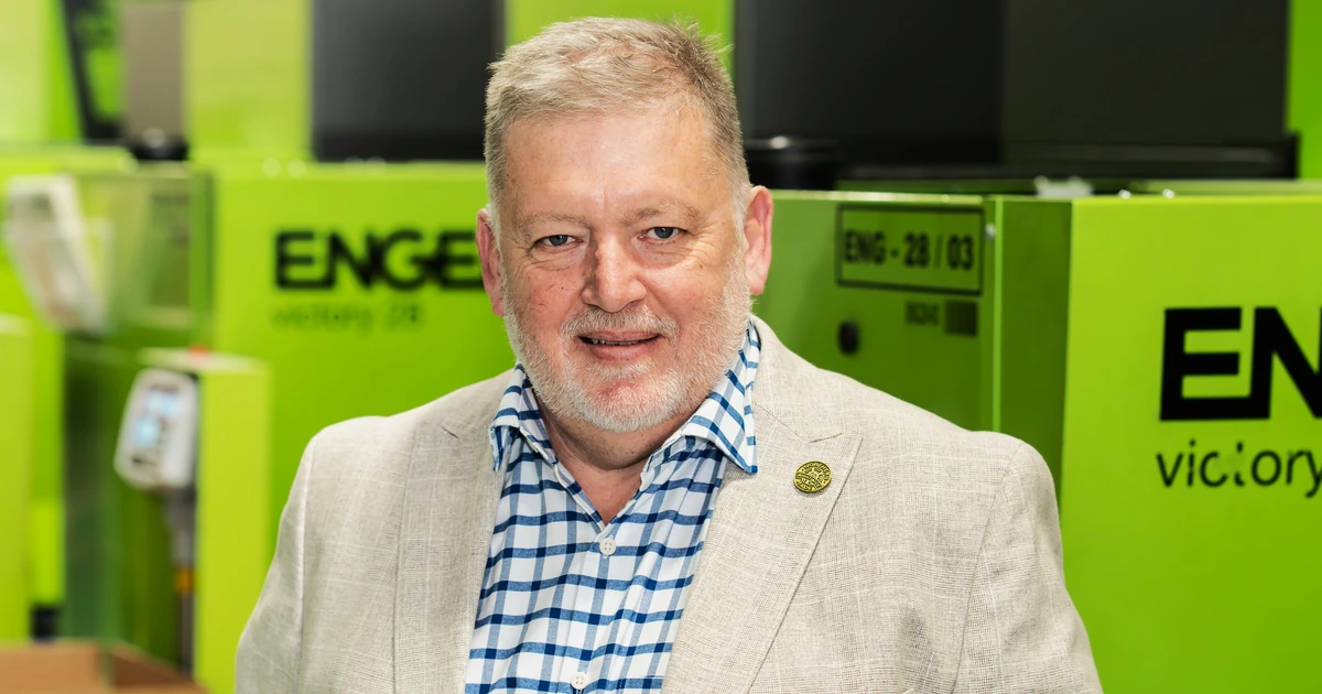 Dean Bradley standing indoors in front of green industrial machines, wearing a checkered shirt and beige jacket