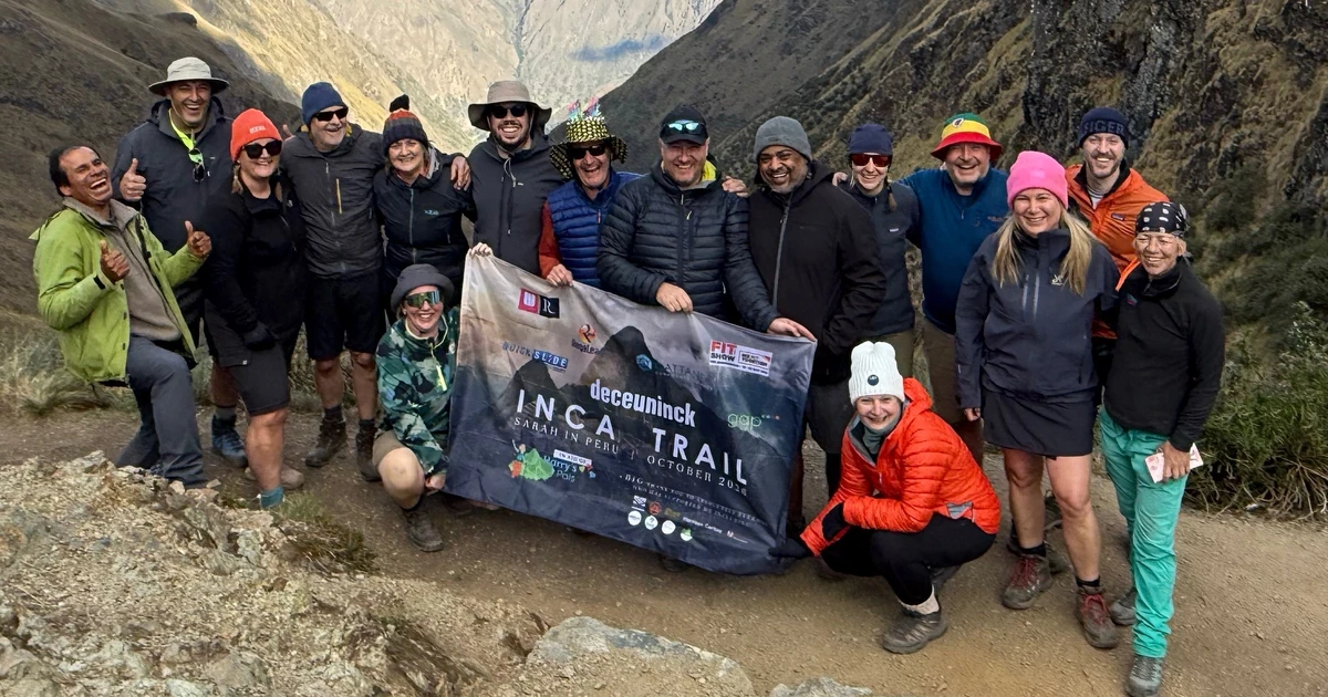 Group of 15 people on mountain trail holding an Inca Trail fundraising banner with mountainous landscape in background