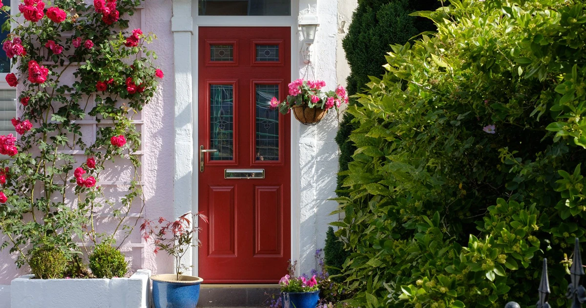 Red front door with decorative glass panels, surrounded by pink flowers and green shrubs at a cottage entrance